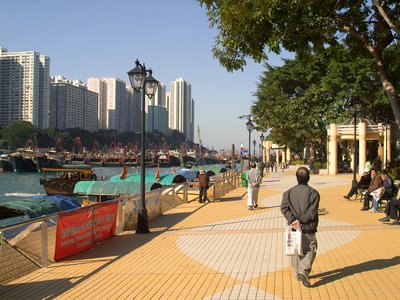 Pedestrians walking down Aberdeen Promenade with boats docked nearby and skyscrapers in distance, Hong Kong island