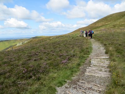 Curved stone pathway leading towards female group of outdoor walkers surrounded by grassy hills and Lake District landscape