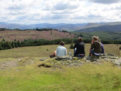 Walking group of women enjoying lunchbreak on a sunny day with a view of heather-covered fields, pine trees, and Lake District fells in the distance