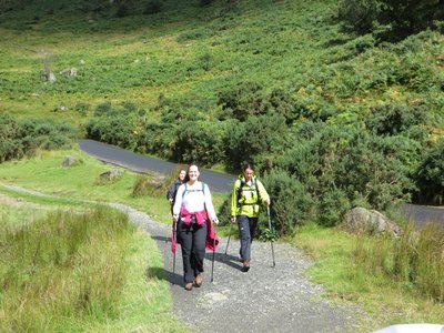 Three women walking towards camera wearing outdoor clothing and using walking poles on stone pathway surrounded by wild shrubs
