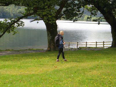 Side-view of woman wearing outdoor clothing and holding walking poles standing on grass with oak trees and Buttermere Lake behind her as she looks into the distance