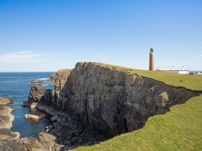 Grassy field leading up to big rocky coastal edge and sea view with Butt of Lewis lighthouse in distance, Scotland