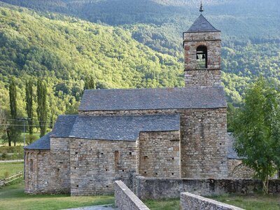 Side view of romaneque church on sunny day with mountainside in background filled with green trees, Vall de Boi, Spain