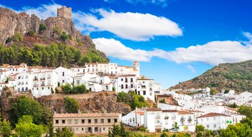 White and beige buildings nestled into hills of mountainous area with tall pine trees dotted throughout and a castle atop the highest point during sunny day, Zahara de la Sierra town, Sierra de Grazalema, Cadiz, Andalusia, Spain