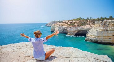 Woman sat on cliff edge with arms spread out enjoying view of turquoise waters and coastal view on coast of western Algarve, Portugal