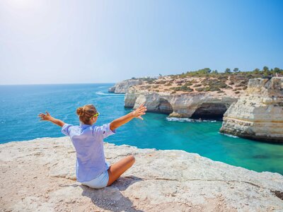 Woman sat on cliff edge with arms spread out enjoying view of turquoise waters and coastal view on coast of western Algarve, Portugal
