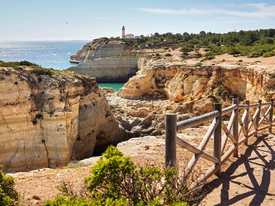 Coastal view of Seven Hanging Valleys walking trail with wooden fenced pathway next to cliffs and lighthouse in distance, Algarve, Portugal