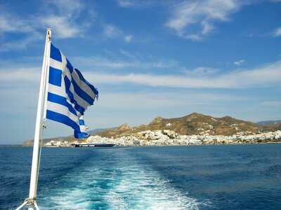 Flag of Greece waving on rear view of boat with white water trail moving toward Greek island in distance, Greece