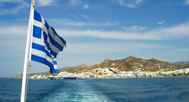Flag of Greece waving on rear view of boat with white water trail moving toward Greek island in distance, Greece