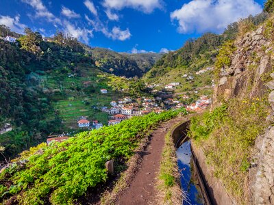 Levada walking trail with irrigation channel to right and madeiran village Marocos in background nestled into mountain side, Portugal