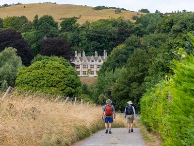 High angle view over downward sloping public footpath Cotswolds Way with two hikers heading downhill between grasses and trees with traditional buildings between green in distance, near Winchcombe, UK