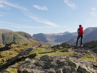 Hiker with bright red rucksack looks out over Buttermere in the Lake District from the top of a hill at Rannerdale Knotts