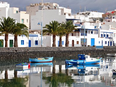 White and blue painted buildings with palm trees by sidewalk and small boats floating in dock nearby, Port city of Arrecife, Lanzarote, Canary Islands, Spain